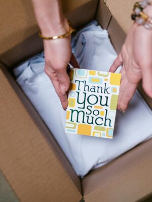 A person holds a thank you card above items in an open shipping box, suggesting care in sending.